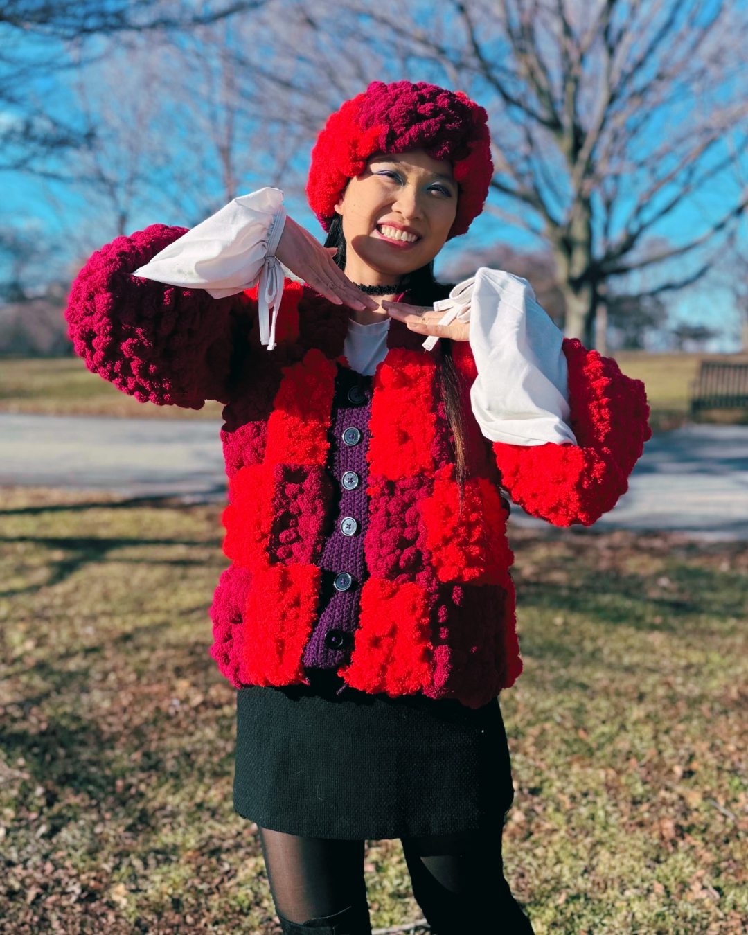 A woman is smiling and posing with hands under chin in a handmade checkered crochet jacket and matching headband